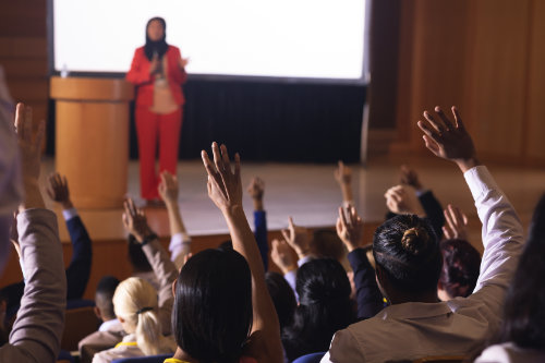 Front view of mixed race businesswoman standing around the podium in the auditorium while audience raising hand for asking question
